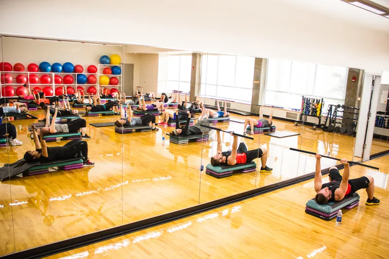 Cours de musculation en groupe dans une grande salle avec miroir : les participants, couchés sur leur step, poussent leur barre vers le haut.