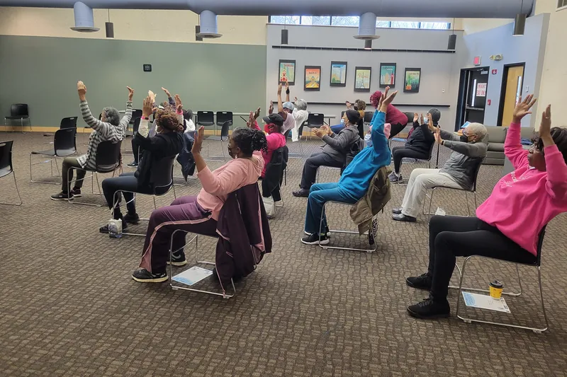 Groupe de personnes aînées assises sur des chaises qui lèvent les bras dans une salle communautaire.