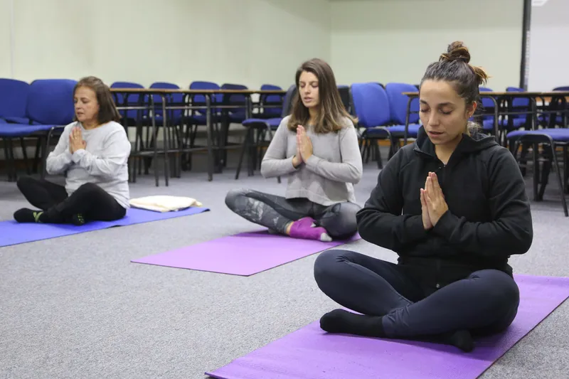 Trois participantes assises en tailleur sur leur tapis, mains jointes et yeux fermés, pendant un moment de méditation.